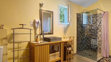 Un baño con tocador de madera, un espejo, un secador de pelo, ducha con puerta de cristal y una ventana pequeña.