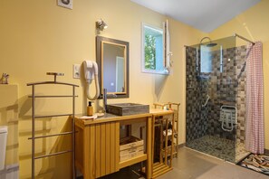 Une salle de bain avec un meuble-lavabo en bois, un miroir, un sèche-cheveux, une douche avec une porte en verre et une petite fenêtre.