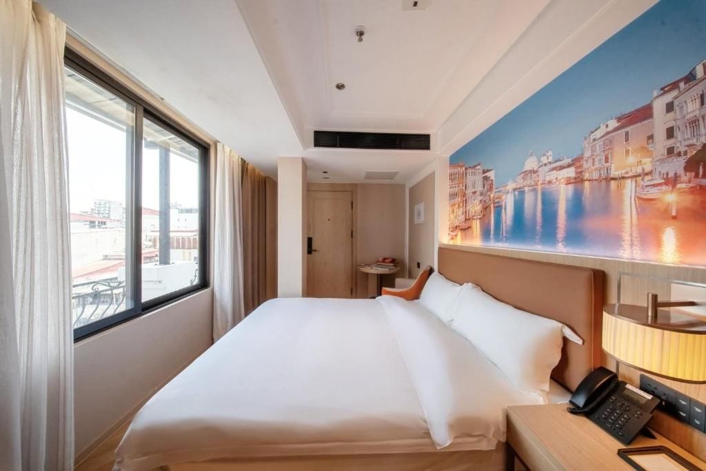 A hotel room with a large bed, a bedside table with a lamp, a painting of a cityscape, and a window with curtains.