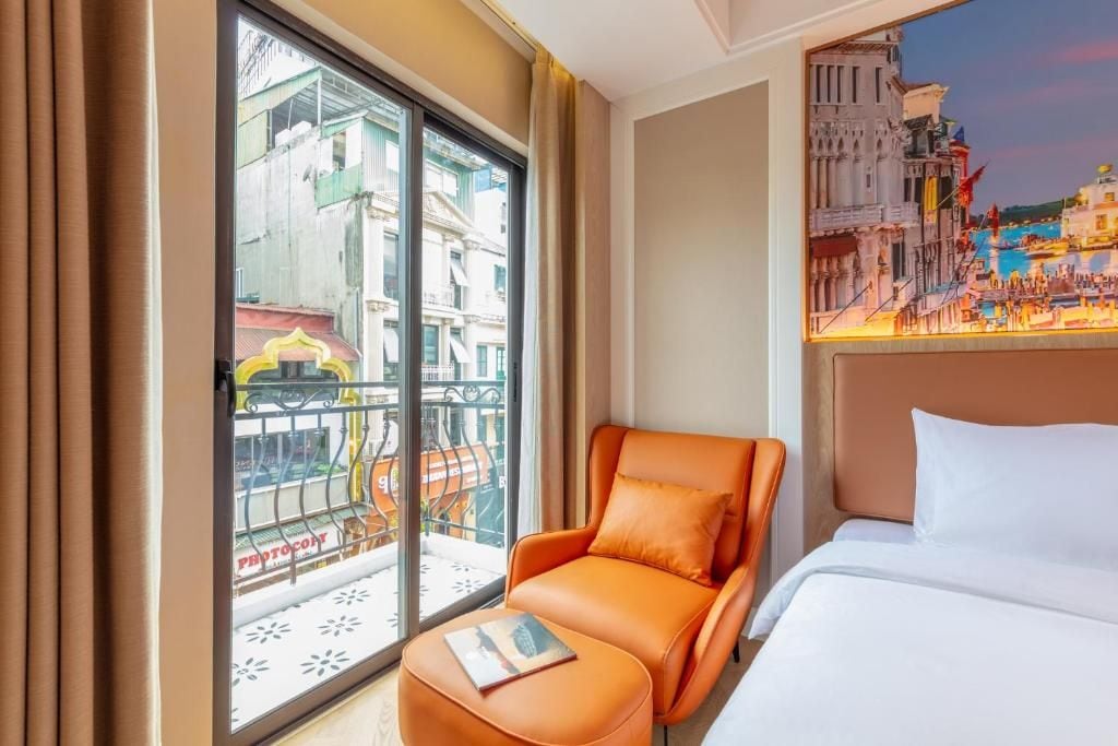 A hotel room with a bed, an orange armchair, a small table, a balcony with a view of buildings, and a painting of a cityscape.
