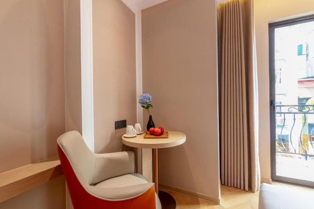 A corner room with a red chair, a small round table with a vase and fruit, and a view through a window with curtains.