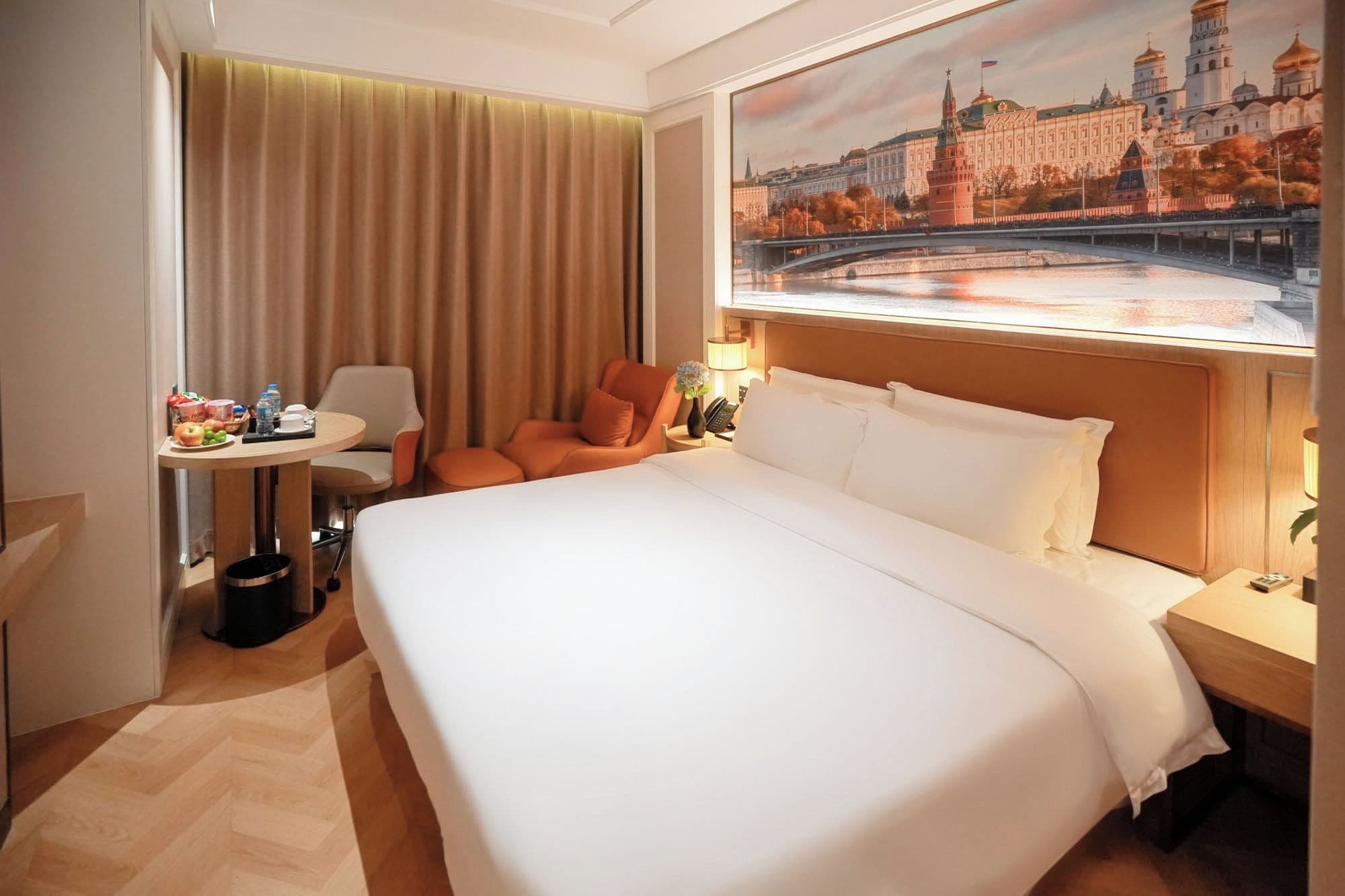 Une chambre d’hôtel avec un grand lit, un bureau avec une chaise, une petite table avec des collations et un tableau représentant un paysage urbain.