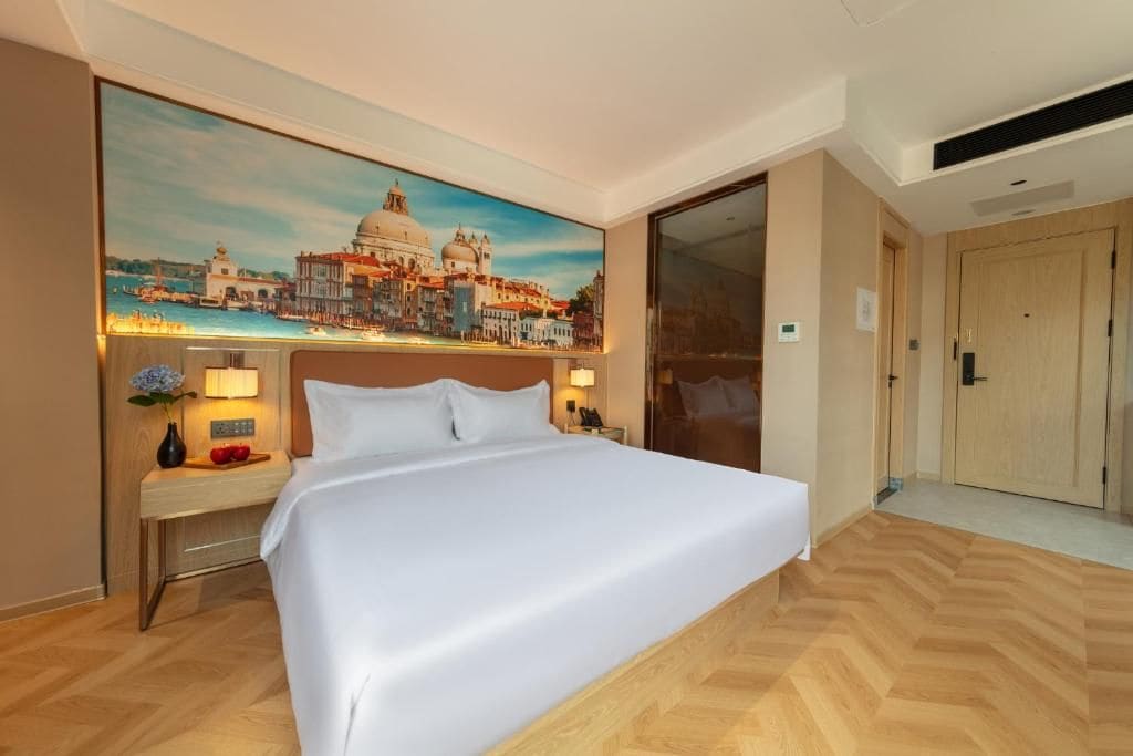 A hotel room with a bed, a nightstand, a lamp, a painting of a cityscape, and a door.