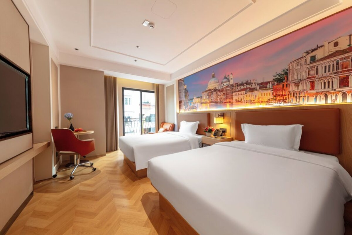 A hotel room with two beds, a desk, a TV, and a large cityscape painting on the wall.