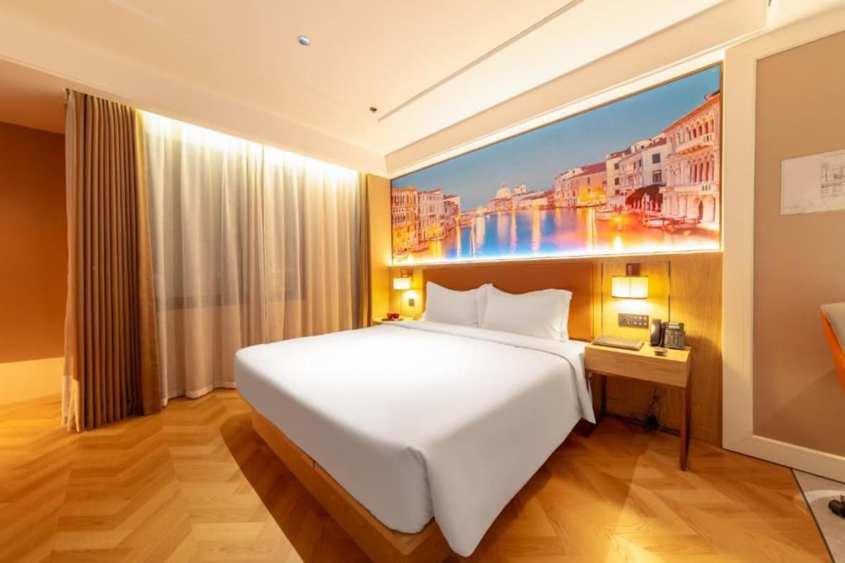 Camera d'albergo con un letto grande, un comodino, una lampada e una TV a muro che mostra una veduta sulla città.