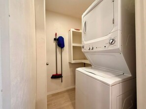 A laundry room with a front-loading washer and dryer, a mop rack, and a broom. - Casa Sol Azul 1A - Cozy 2BR, Ground Floor, Pool! (South Padre Island)