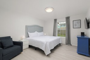 A bedroom with a bed, a bedside table, a lamp, a chair, a window with curtains, and a blue cabinet. - Casa Sol Azul 1A - Cozy 2BR, Ground Floor, Pool! (South Padre Island)