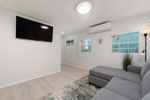 Ein Wohnzimmer mit einem an der Wand befestigten Flachbildfernseher, einem grauen Sofa, einem gemusterten Teppich und einem Fenster mit Jalousien.