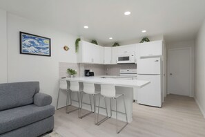 Eine moderne Küche mit weißen Schränken, einer weißen Arbeitsplatte und Barhocker. Ein Gemälde hängt an der Wand, und ein graues Sofa befindet sich im Vordergrund.