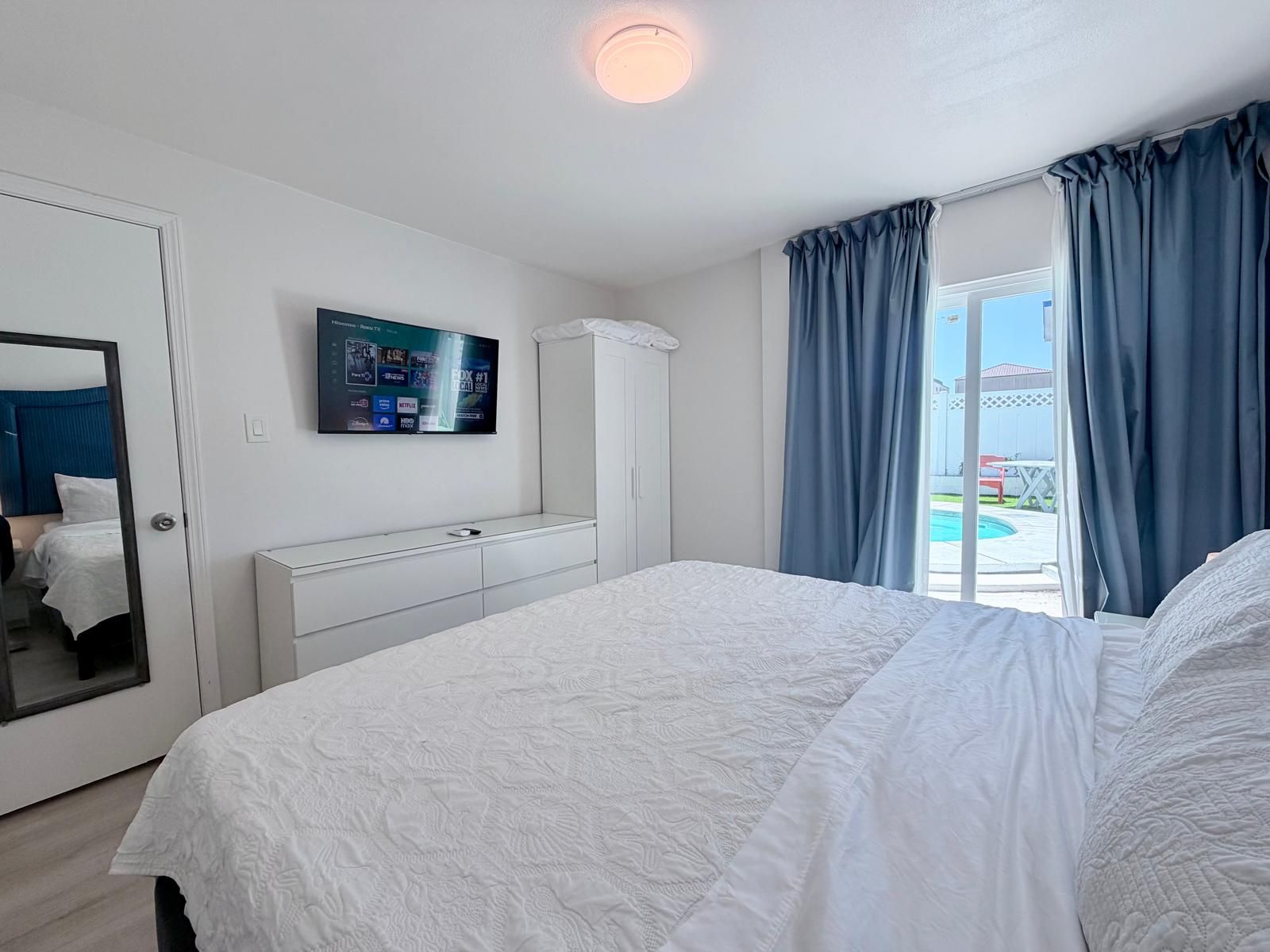 A hotel room with a bed, a TV mounted on the wall, a wardrobe, and a view of a swimming pool.