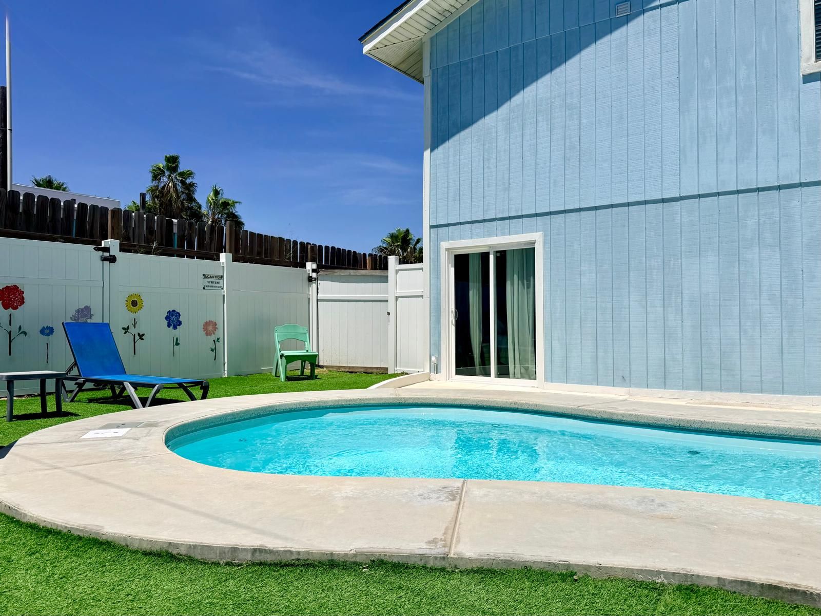 Ein Garten mit Swimmingpool, blauem Stuhl, grünem Stuhl und weißem Zaun mit bemalten Blumen.
