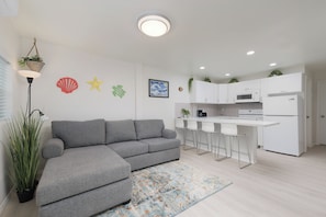 Ein modernes Wohnzimmer mit einem grauen Sofa, eine Küche mit weißen Schränken und einem weißen Kühlschrank sowie ein kleiner Essbereich mit weißen Barhocker.