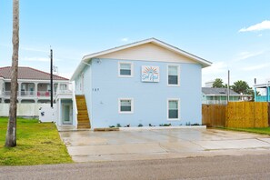 A two-story building with a blue exterior, a wooden front porch, and a small front yard. - Casa Sol Azul 2A - Spacious 2BR, Pool! (South Padre Island)