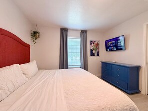 A bedroom with a bed, a blue dresser, a TV on the wall, and a window with curtains. - Casa Sol Azul 2A - Spacious 2BR, Pool! (South Padre Island)