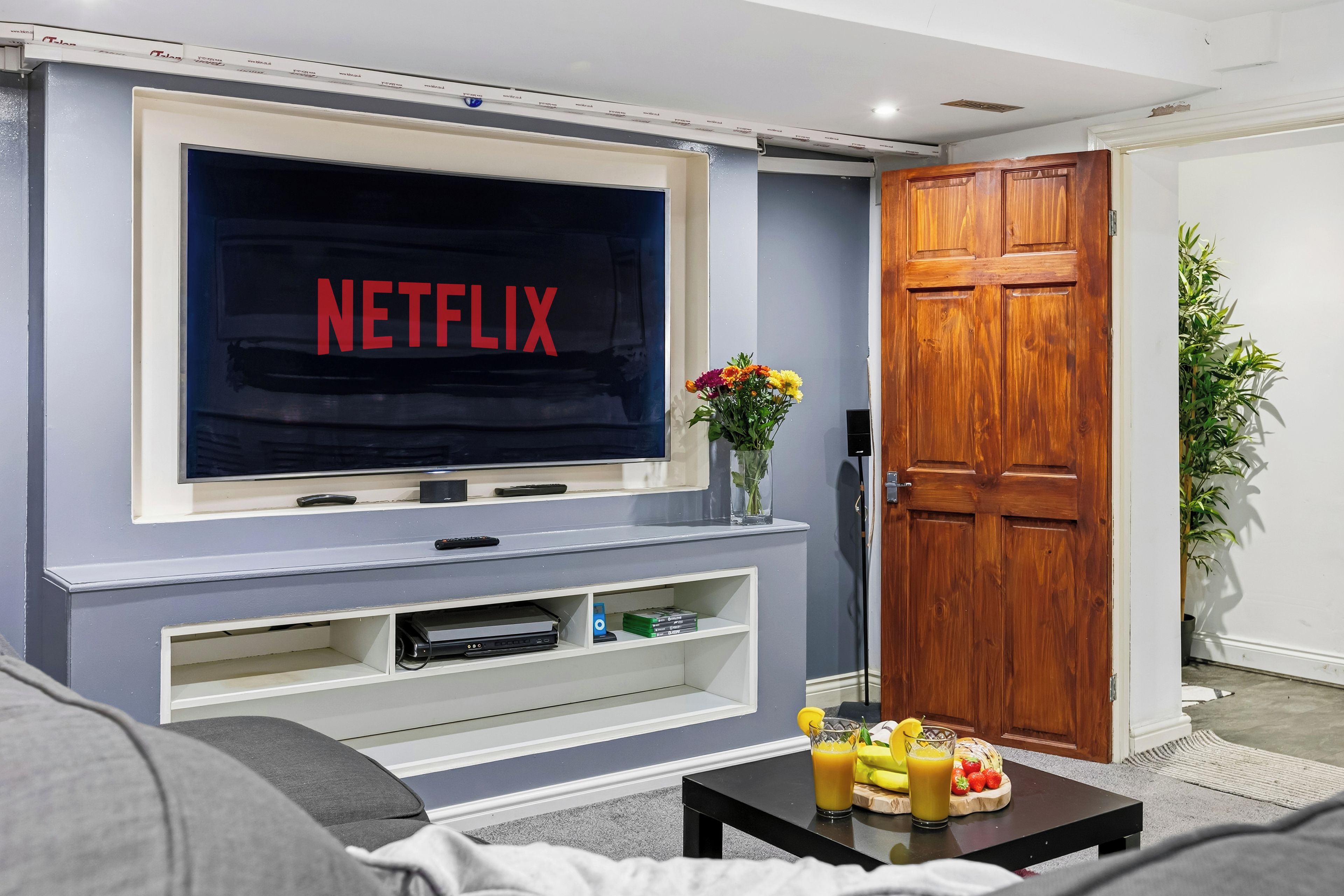 Ein Wohnzimmer mit einem Flachbildfernseher, der 'NETFLIX' zeigt, einer Holztür und einem kleinen Tisch mit Obst und einem Getränk.