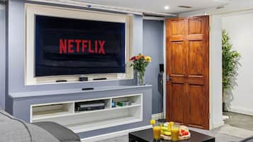 A living room with a flat-screen TV displaying 'NETFLIX,' a wooden door, and a small table with fruit and a drink.