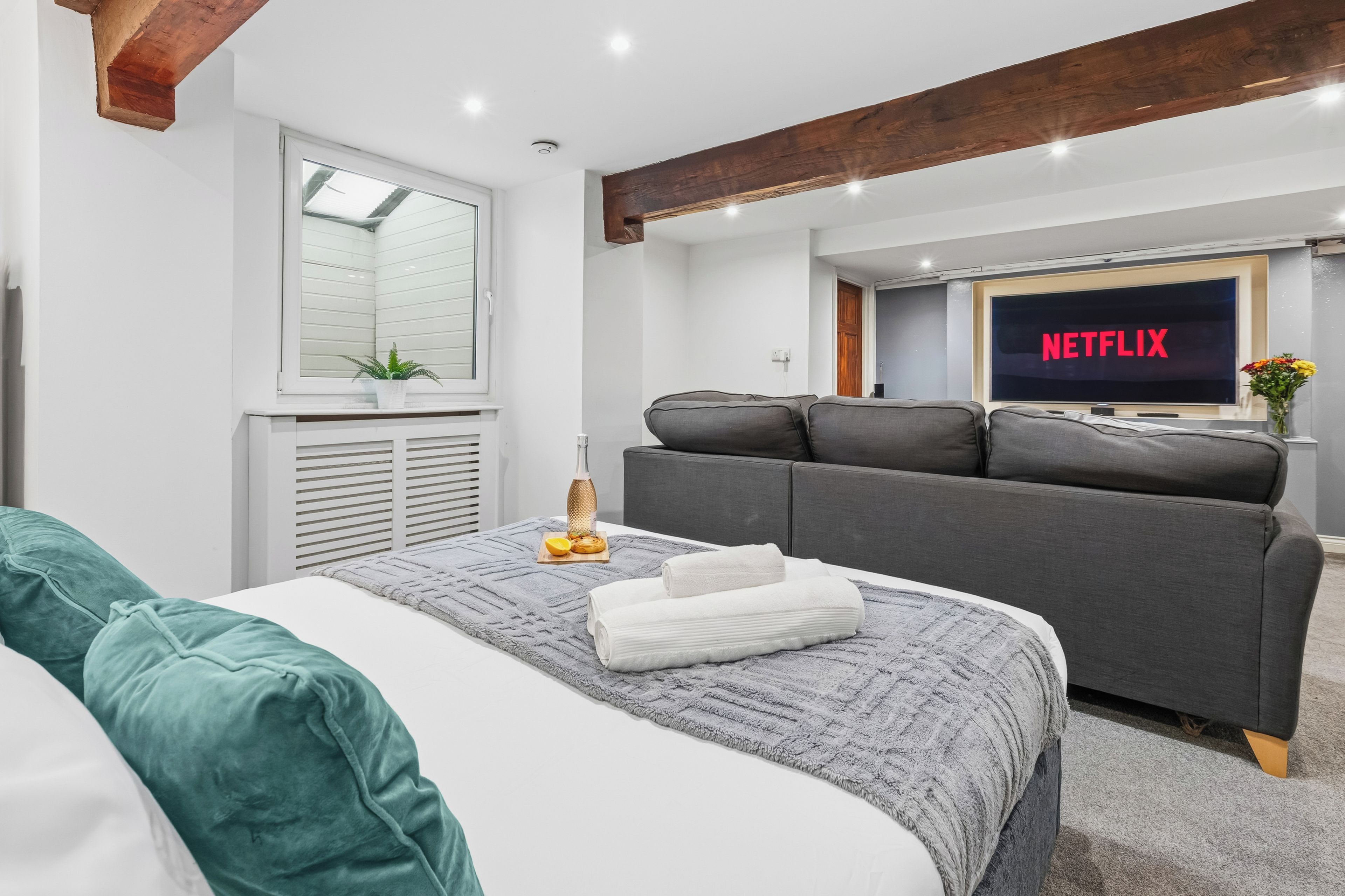Una camera da letto moderna con un letto, un divano grigio e una televisione che mostra 'NETFLIX'.