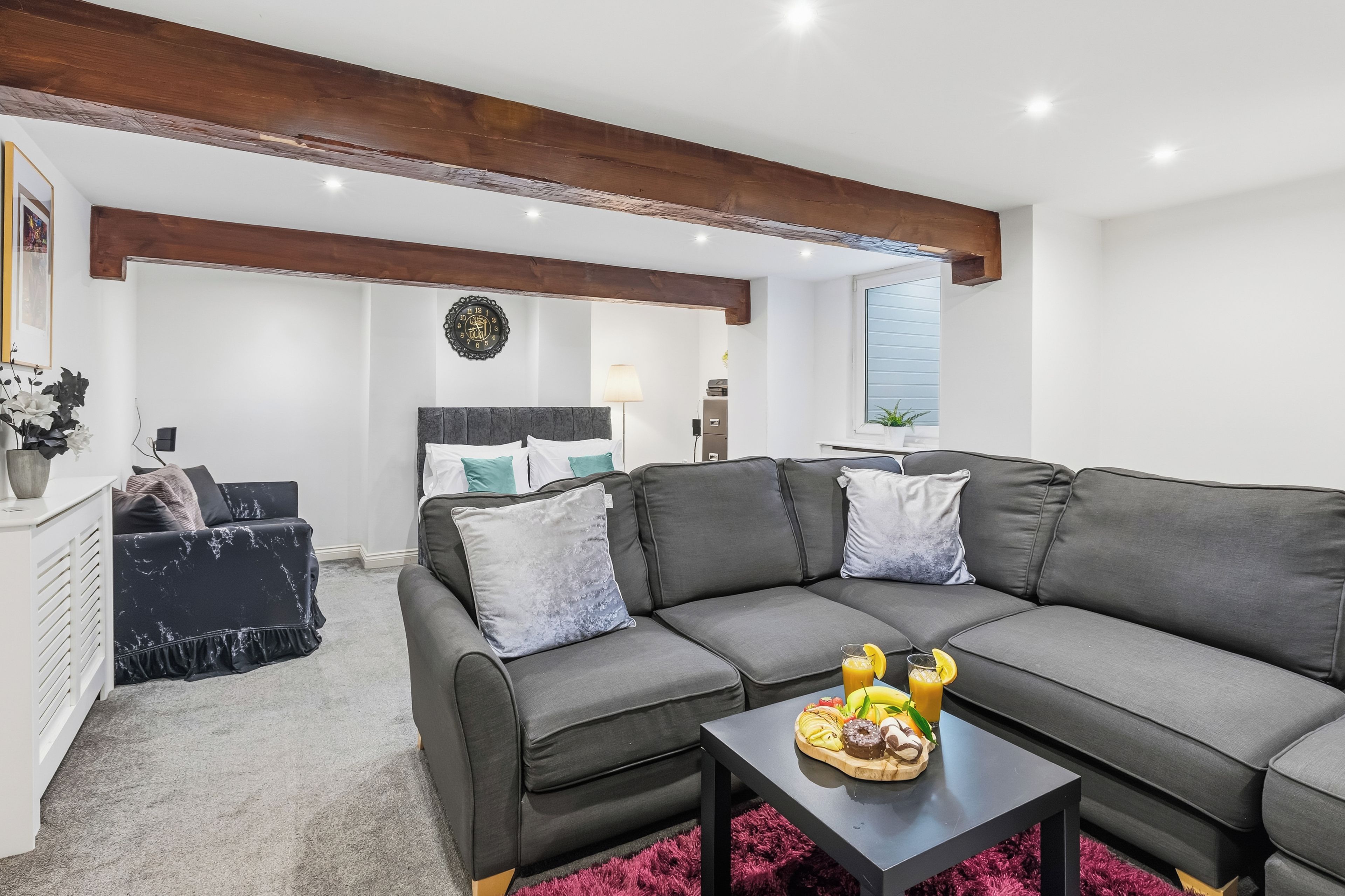 A modern living room with a sectional sofa, a coffee table with fruit and a drink, and a bed in the background.