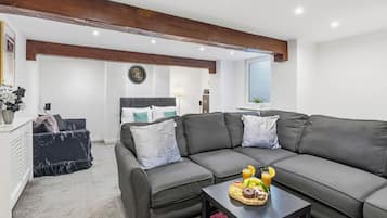 A modern living room with a sectional sofa, a coffee table with fruit and a drink, and a bed in the background.