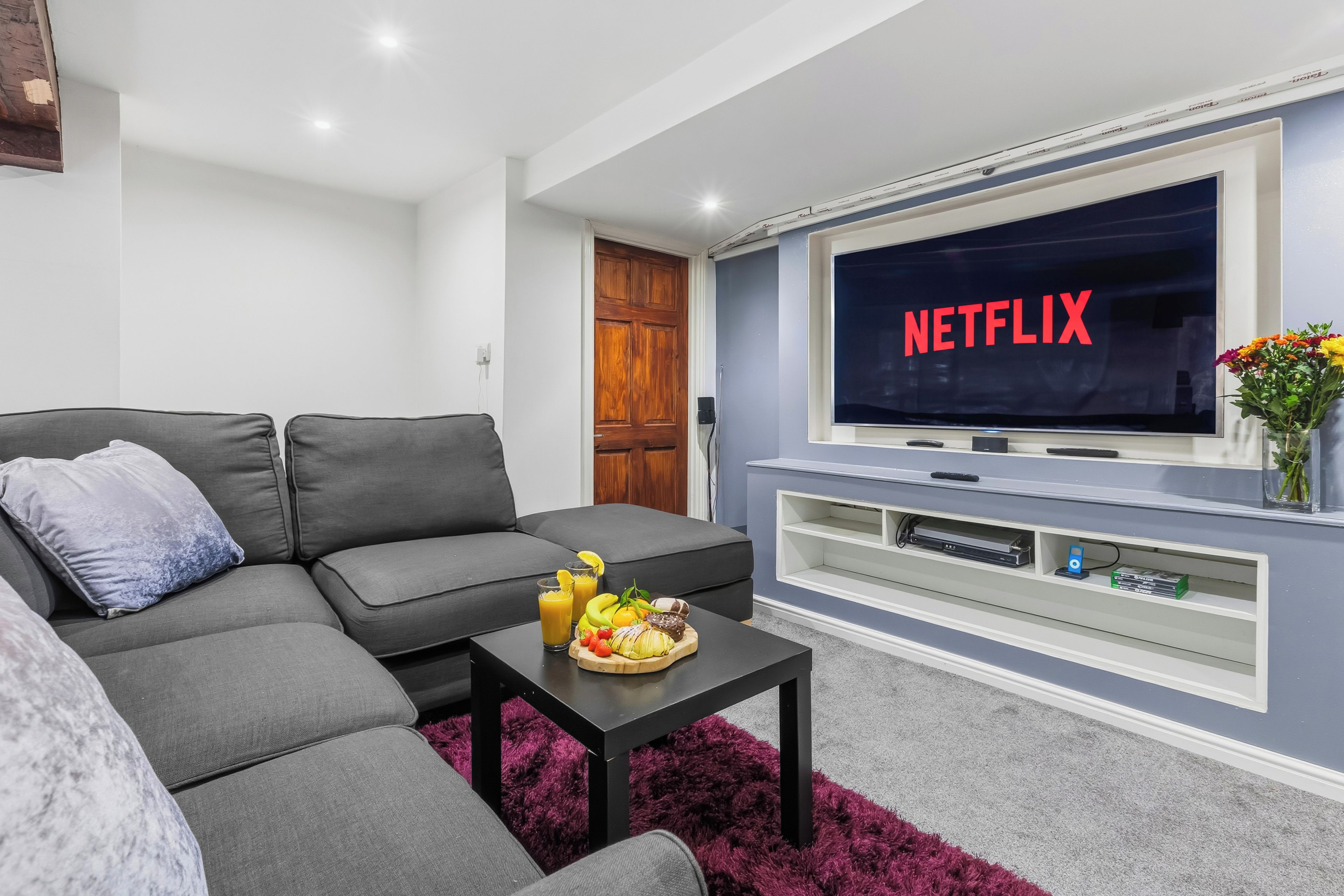 A living room with a grey sofa, a coffee table with fruit, and a TV mounted on the wall displaying 'NETFLIX'.
