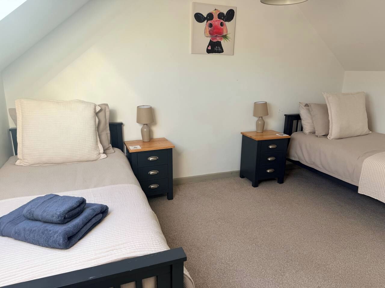 A bedroom with two beds, each with a nightstand, a lamp, and a wall-mounted painting.