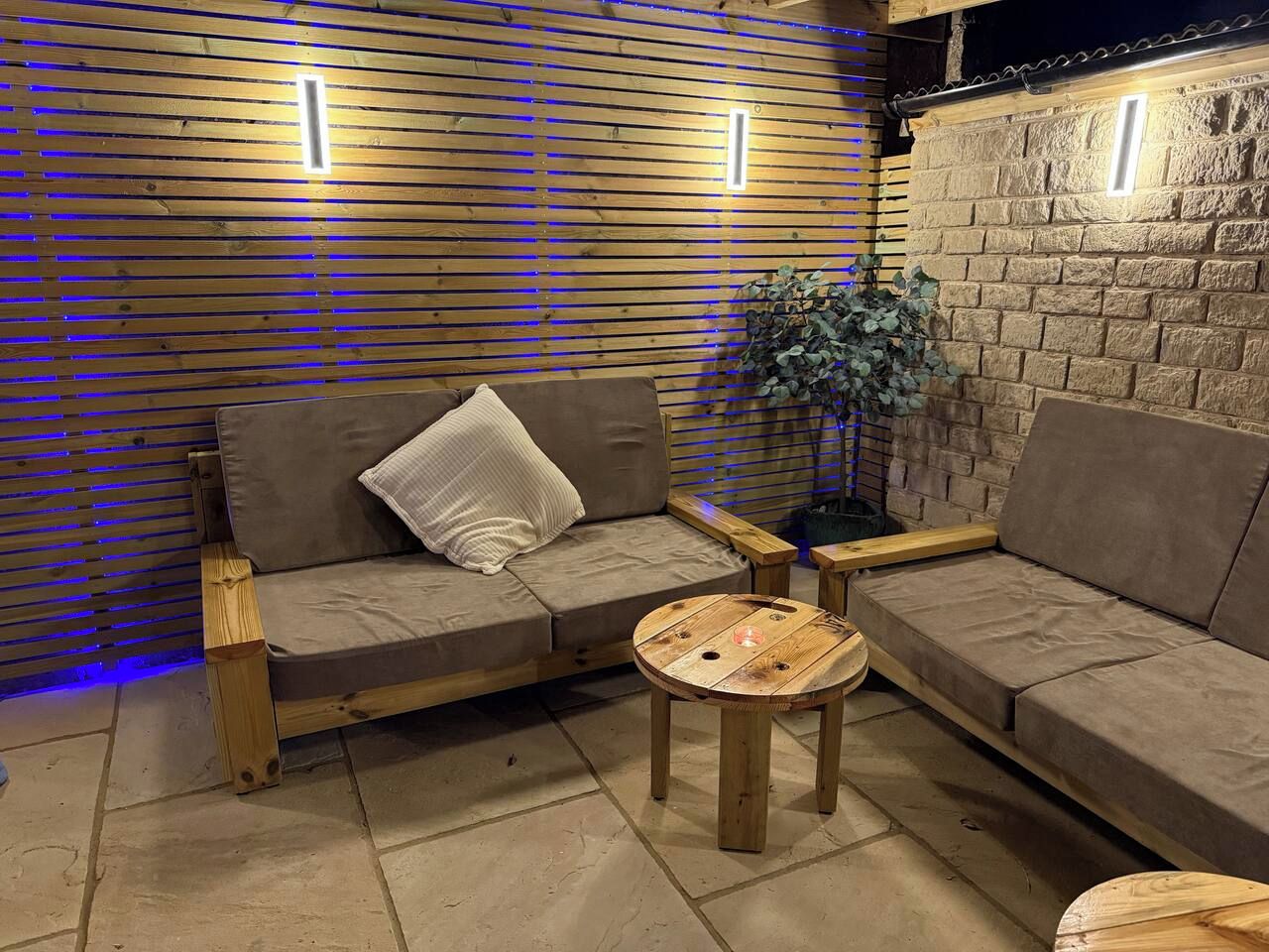 A wooden sofa with cushions, a small round wooden table, and a potted plant in a room with a tiled floor and wooden slatted walls.