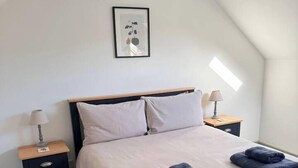 Una cama bien tendida con cabecera, dos mesitas de noche con lámparas y un cuadro enmarcado en la pared.