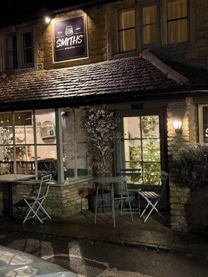 A stone building with a sign reading 'SMITHS OF BOUSTON' and outdoor seating with chairs and tables. - Maple Cottage (Bourton-on-the-Water)