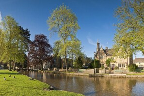 A riverside view with a historic building, a grassy area, and trees. - Maple Cottage (Bourton-on-the-Water)