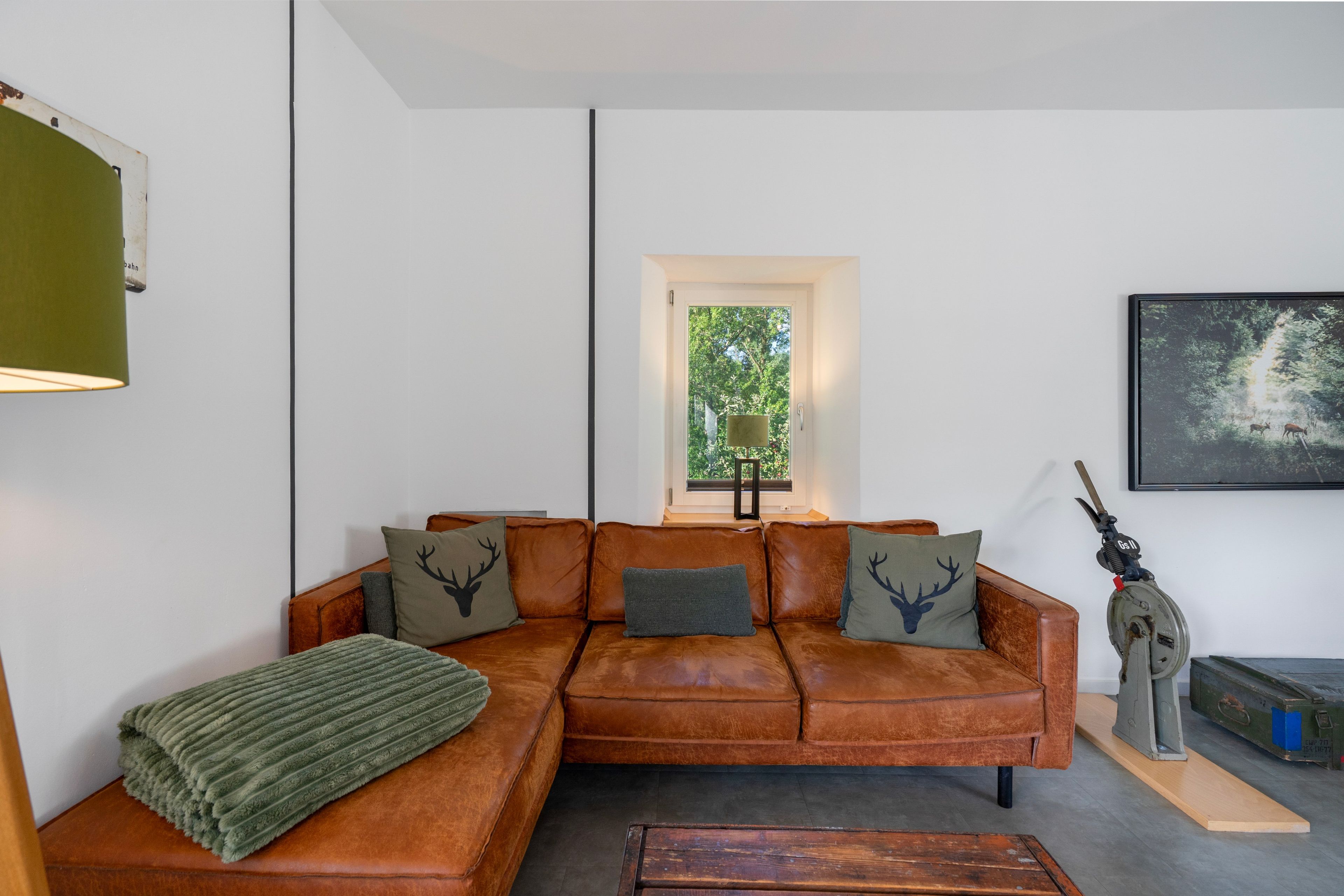 Ein Wohnzimmer mit braunem Ledersofa, einem grünen Überwurf und einem Fenster mit Blick ins Grüne.
