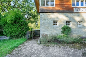 A stone building with a wooden upper section, a metal gate, and a paved path leading to a wooden bench.