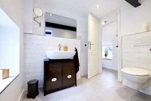 A modern bathroom with a white sink, mirror, and toilet. There is a black cabinet with two doors, a black trash bin, and a window with a plant. - Stellwerk Ringelstein: Forest-Edge Retreat with Sauna, Fireplace, Terrace & Wi-Fi (Büren)