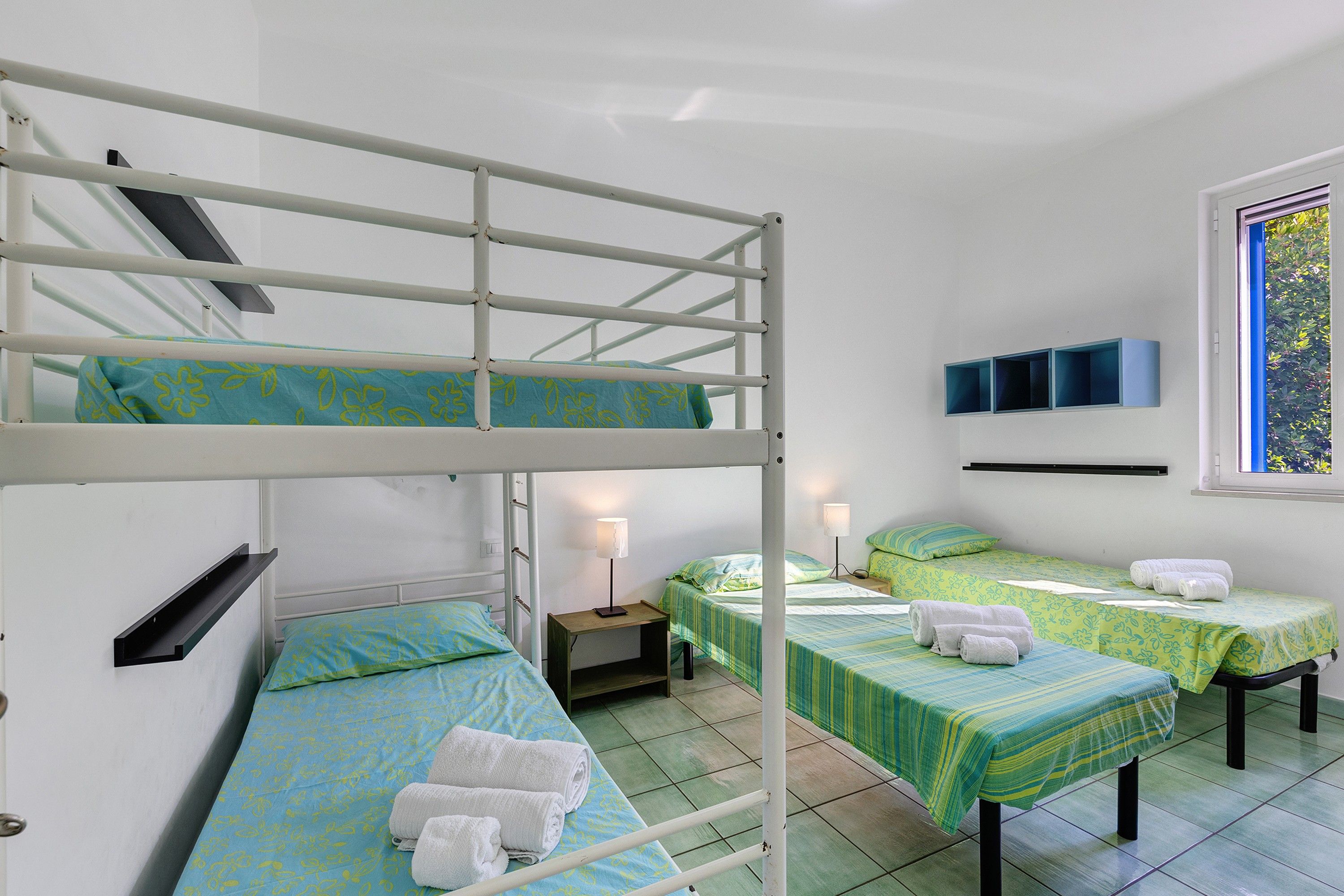 A dormitory room with bunk beds, two single beds, a window with a view, and a small table with a lamp.