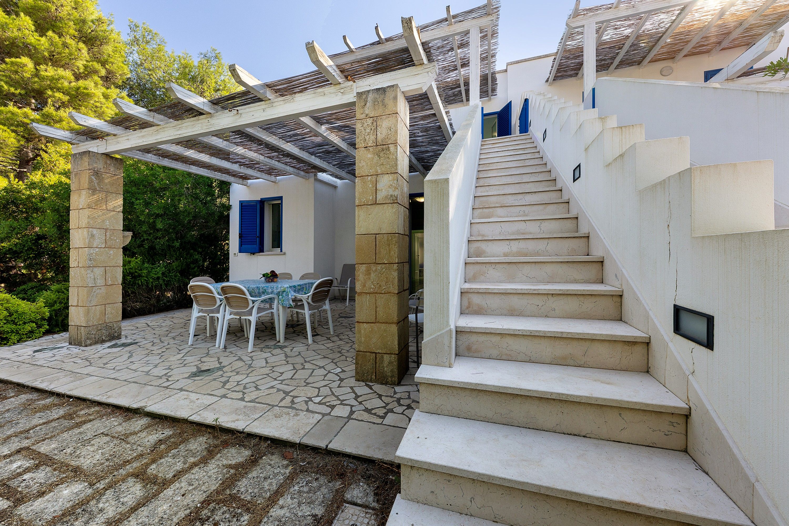 A stone staircase leading to a white building with a blue window, surrounded by greenery.