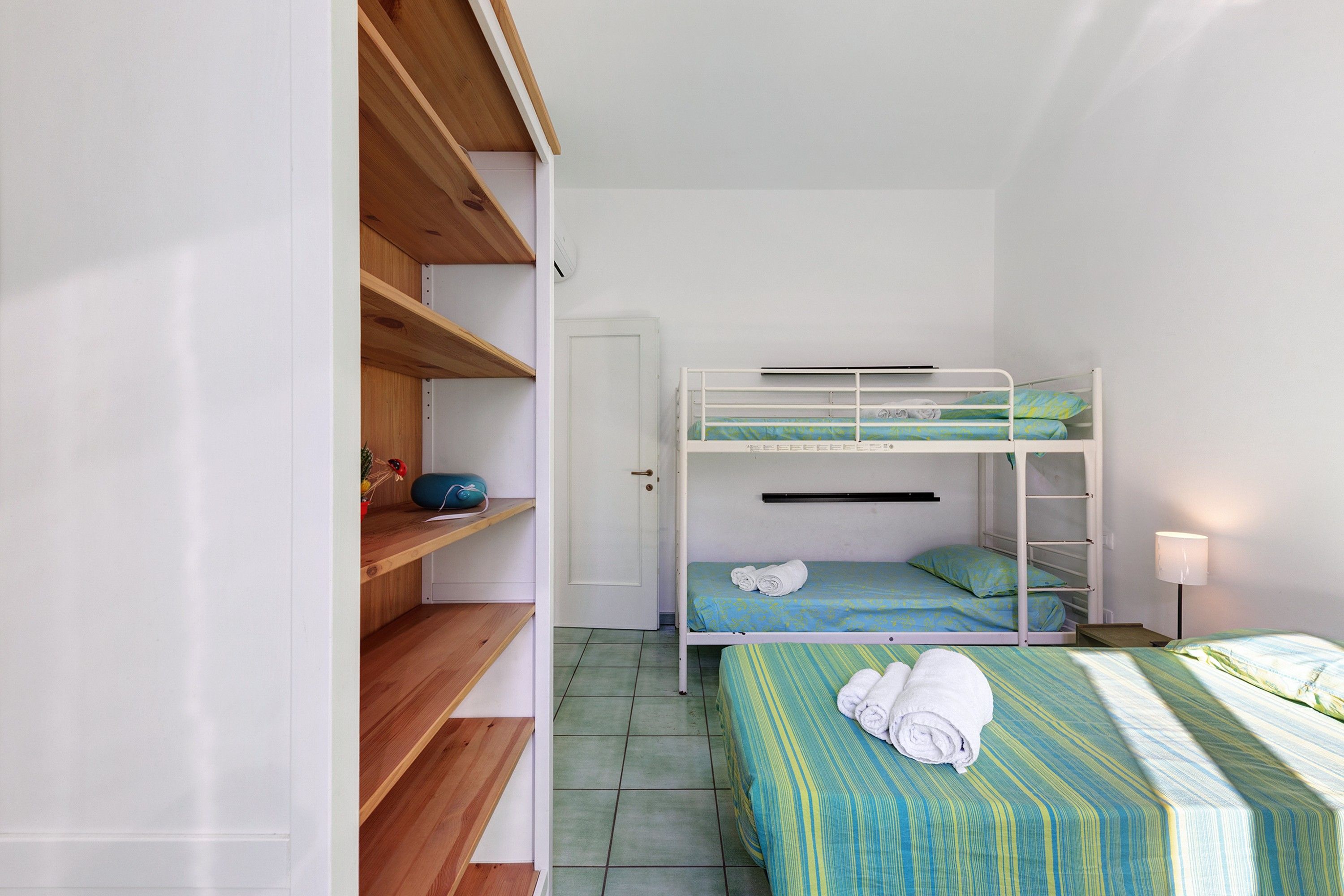 A dormitory room with bunk beds, wooden shelves, a door, and a bedside lamp.