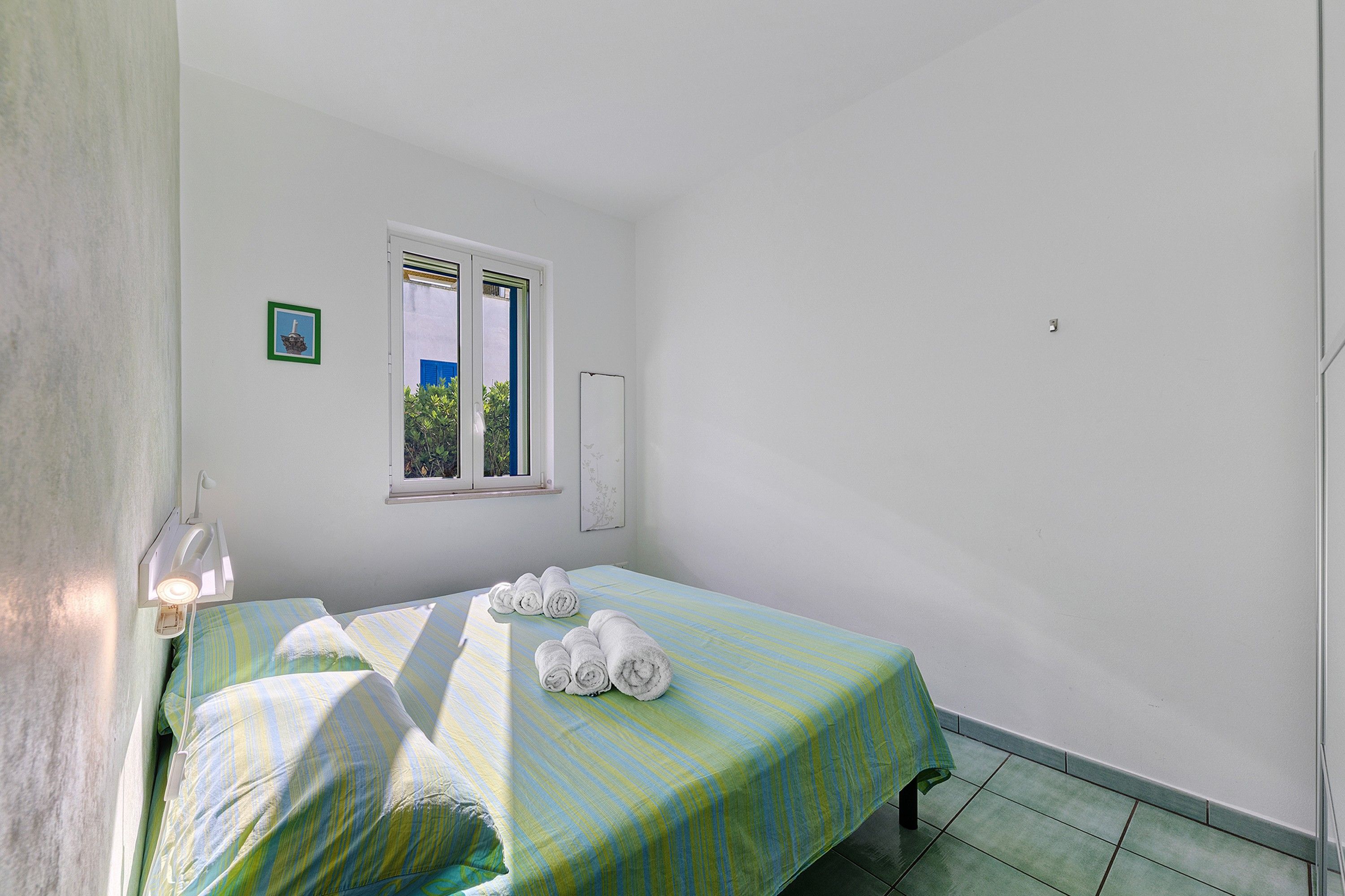 A small, clean room with a single bed, a window, and a wall-mounted light fixture.