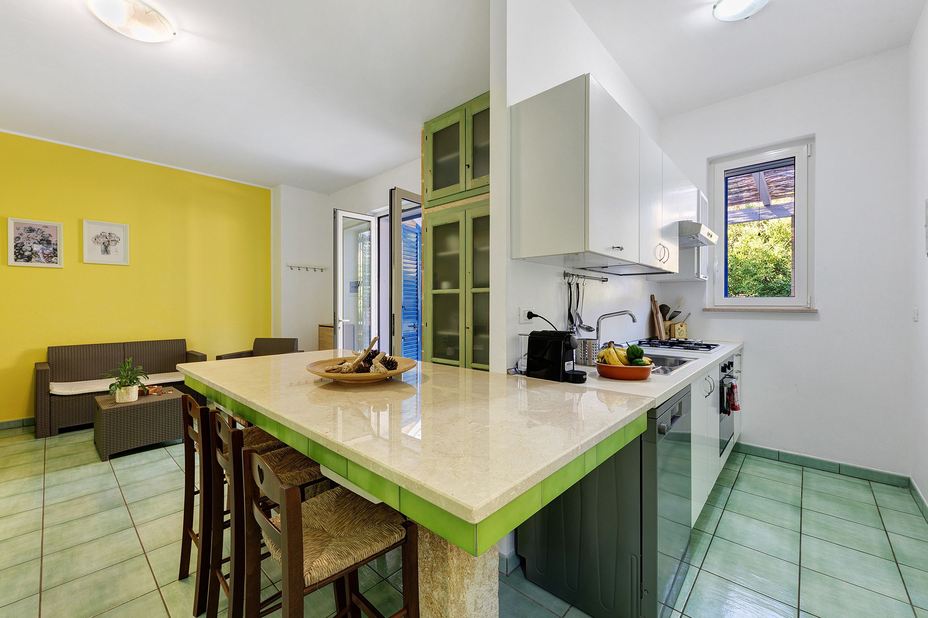 A compact kitchen with a central island, a dining area with a sofa, and a window with a view of greenery.