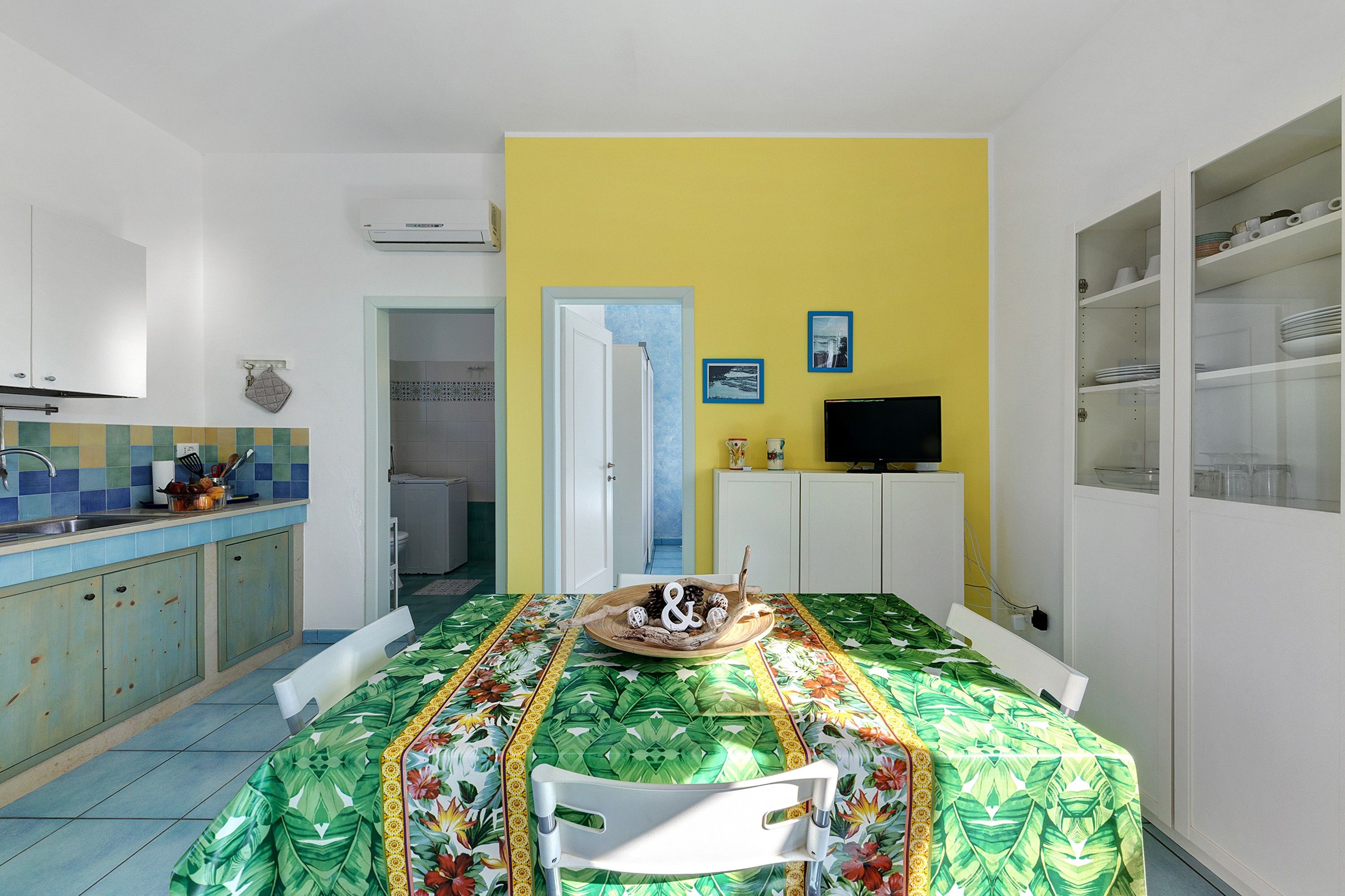 Cocina compacta con salpicadero de azulejos, comedor con mesa para dos personas y un televisor sobre un mueble.