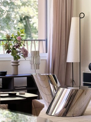 A modern interior with a reflective metallic table, a vase with flowers, a lamp, and a view of greenery outside. - Serenity77 · 100m² designed by an architect, Geneva (Vandœuvres)