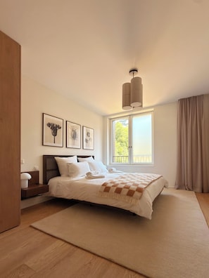 A bedroom with a bed, a window with curtains, a bedside table, and wall art. - Serenity77 · 100m² designed by an architect, Geneva (Vandœuvres)