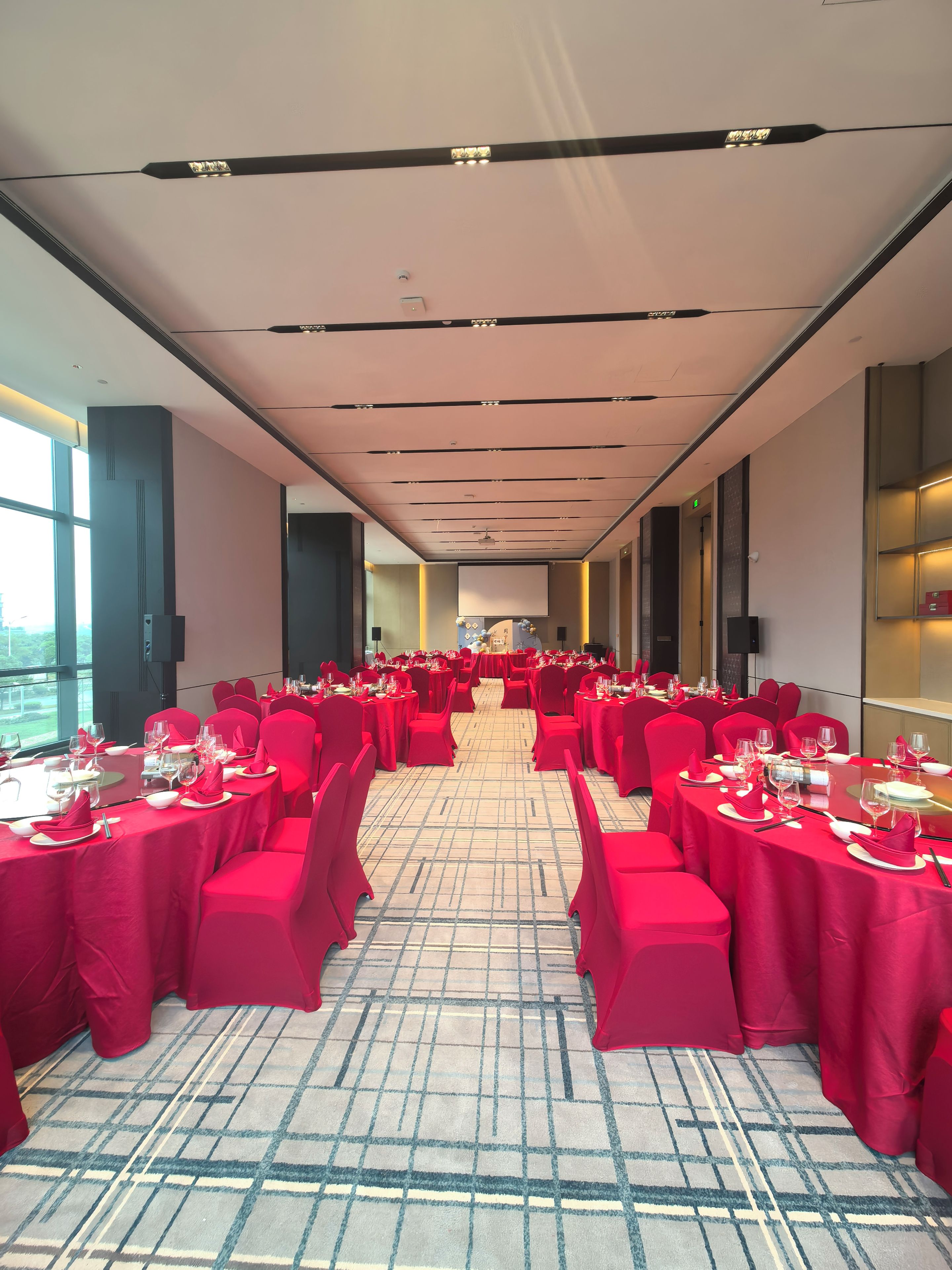 A banquet hall with tables set for dining, a large screen at the front, and a high ceiling with recessed lighting.