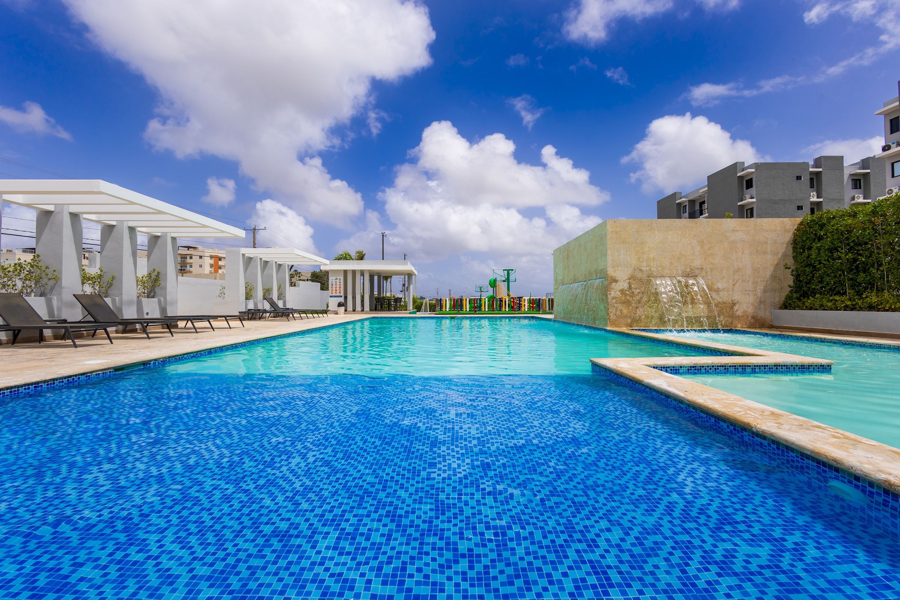 A swimming pool with a tiled deck, lounge chairs, and a covered seating area.