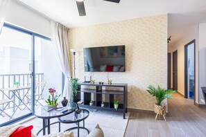 Ein modernes Wohnzimmer mit einem an der Wand befestigten Flachbildfernseher, einem Glastisch und einem Sofa mit einem roten Kissen. Ein Balkon bietet einen Ausblick ins Freie.