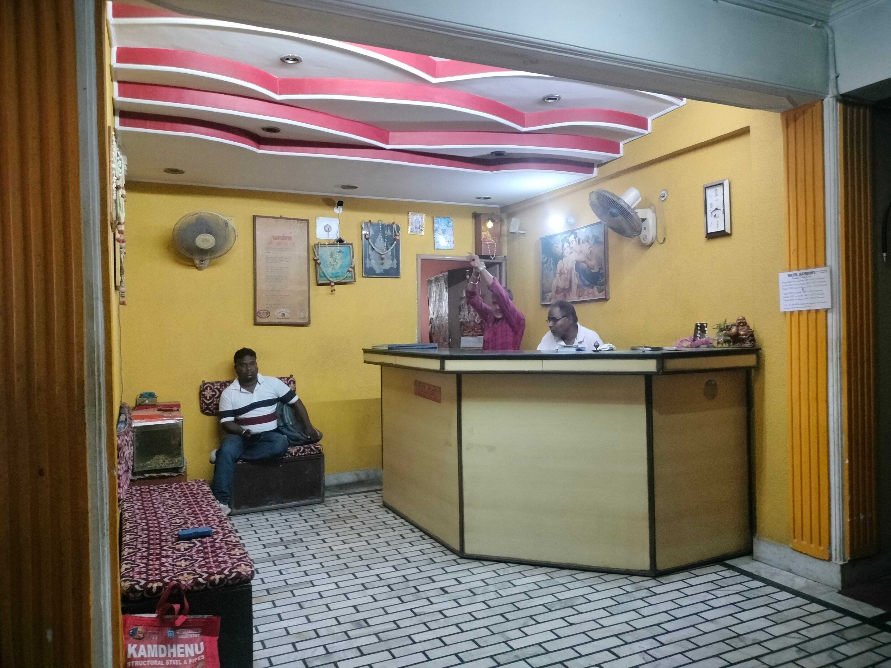 Zona de recepción con mostrador, un huésped sentado, y una pared con cuadros enmarcados y un ventilador.