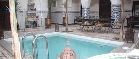 A pool area with a tiled floor, a pool with a decorative fountain, and a table with chairs.