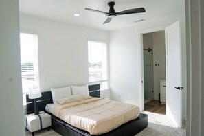 Un dormitorio con cama, mesitas de noche, ventilador de techo y un baño visible a través de una puerta abierta.