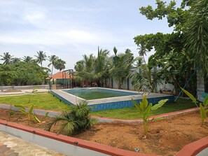 A swimming pool surrounded by a garden with palm trees and a small building in the background. - Villa Nova Resort (Puducherry)