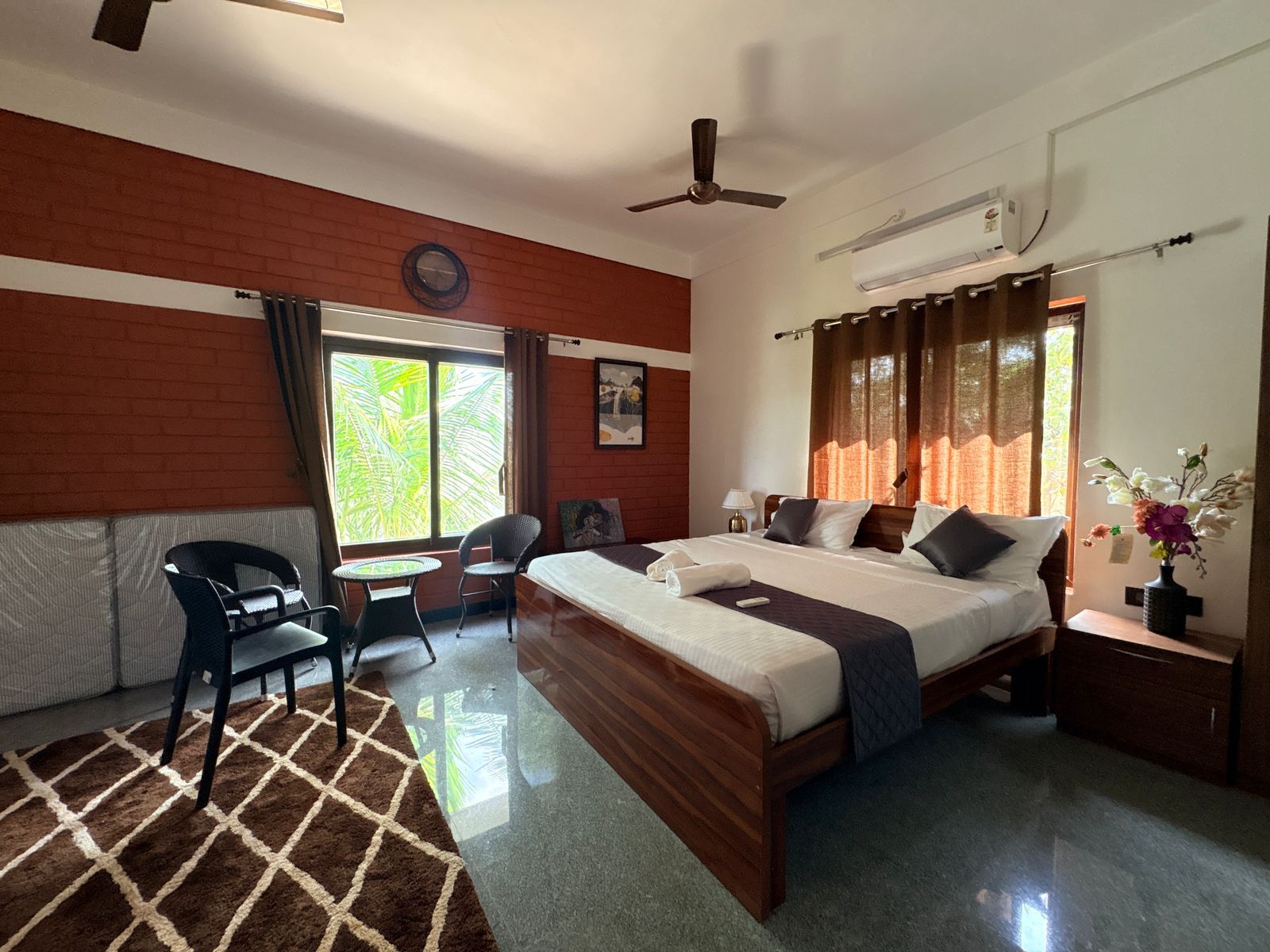 Quarto de hotel com cama, escrivaninha, cadeira, janela com cortinas, ventilador de teto e relógio de parede.