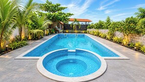 Una piscina rectangular con un jacuzzi circular en el centro, rodeada por una terraza empedrada y abundante vegetación.