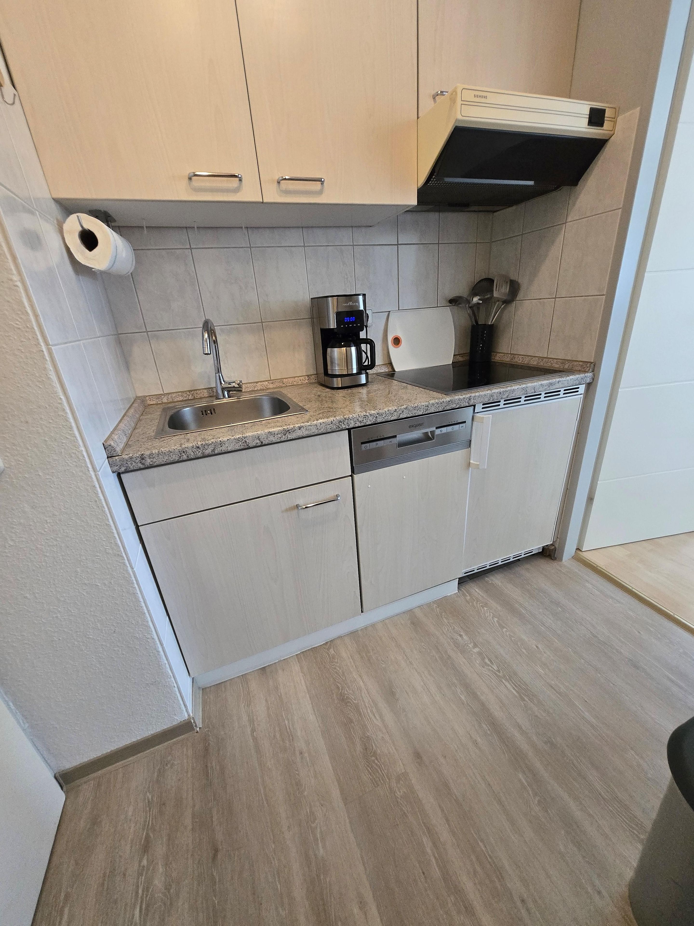 A kitchen with a coffee maker, a dishwasher, and a roll of toilet paper on the wall.